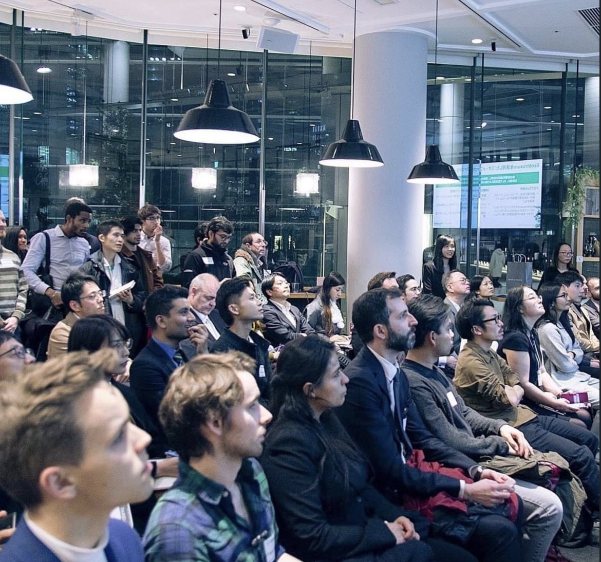A packed audience in a modern glass-walled venue with black pendant lamps overhead, facing a presenter off-frame; the front rows are a mix of researchers, funders and practitioners of many nationalities, and a bilingual Japanese-English programme is visible on a screen at the back.