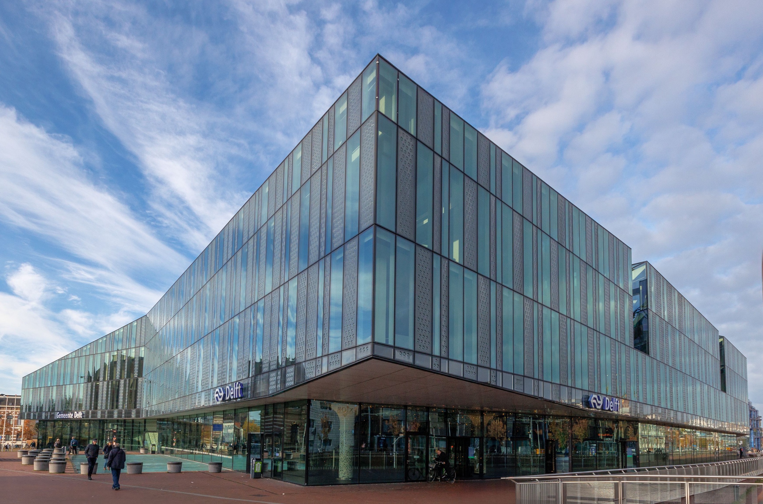 Delft station and Gemeente Delft building on a bright day: a modern glass structure with vertical fritted panels reflecting the sky, low canopies over the entrances and a paved plaza in front.