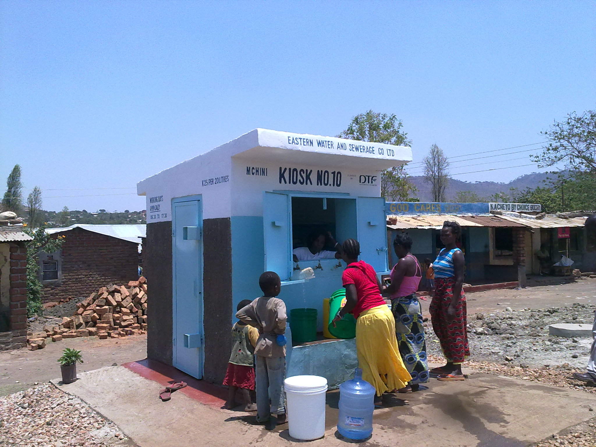 A small blue-and-white metered water kiosk with a chalked tariff and a queue of women and children filling green and yellow jerry cans at the counter window.