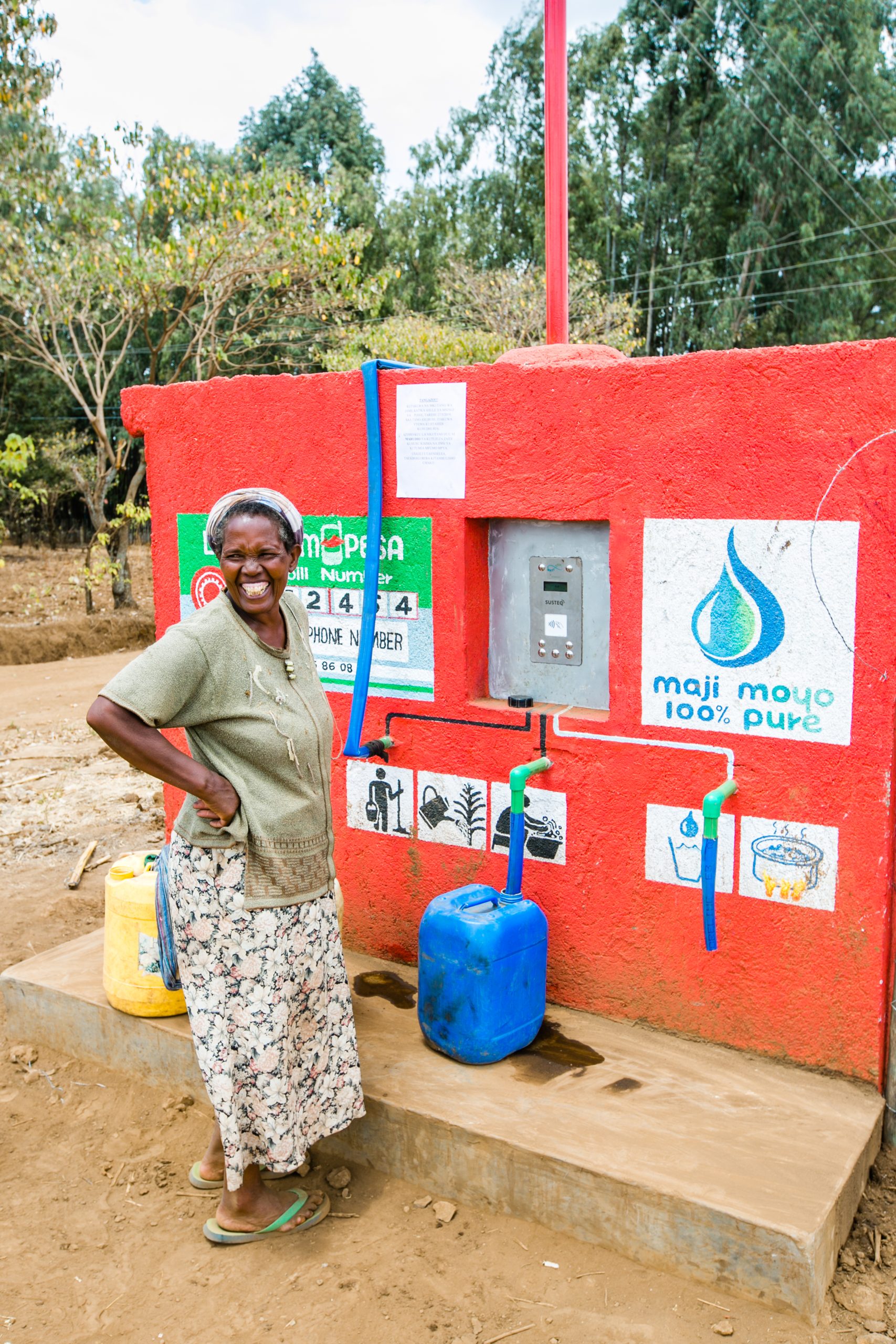 A woman in a floral skirt laughs beside a bright red Maji Moyo vendor kiosk with an M-PESA paybill number and two green taps; a yellow and a blue jerry can at her feet.