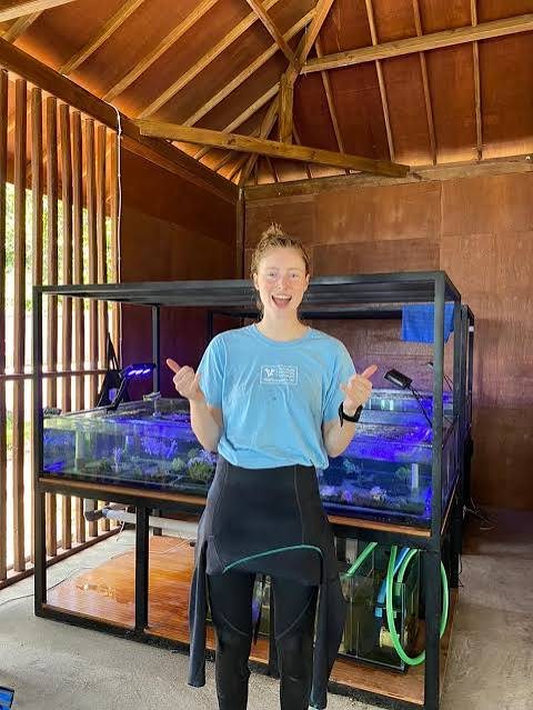 Amba Stapert giving a two-thumbs-up in front of the coral display tanks at the Lombok lab, wearing a light-blue Reef Support t-shirt with a wetsuit pulled down to the waist.