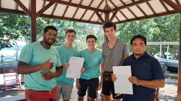Five people standing under a Lombok beach gazebo, holding signed agreements between them; palms and the water visible in the background.