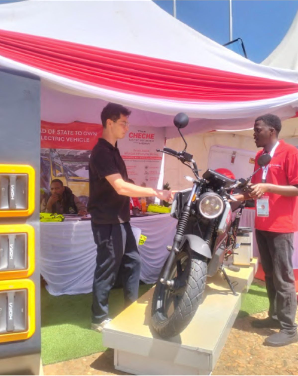 A Cheche electric-motorbike trade-show booth under a red-and-white striped canopy; a young man in a black polo shirt hands a leaflet to a booth attendant in a red Cheche polo standing behind a matte-grey electric motorcycle on a raised white platform; a Head of State to Own Electric Vehicle banner behind the bike; a stack of yellow battery-swap lockers to the left; an attendee seated behind the counter.