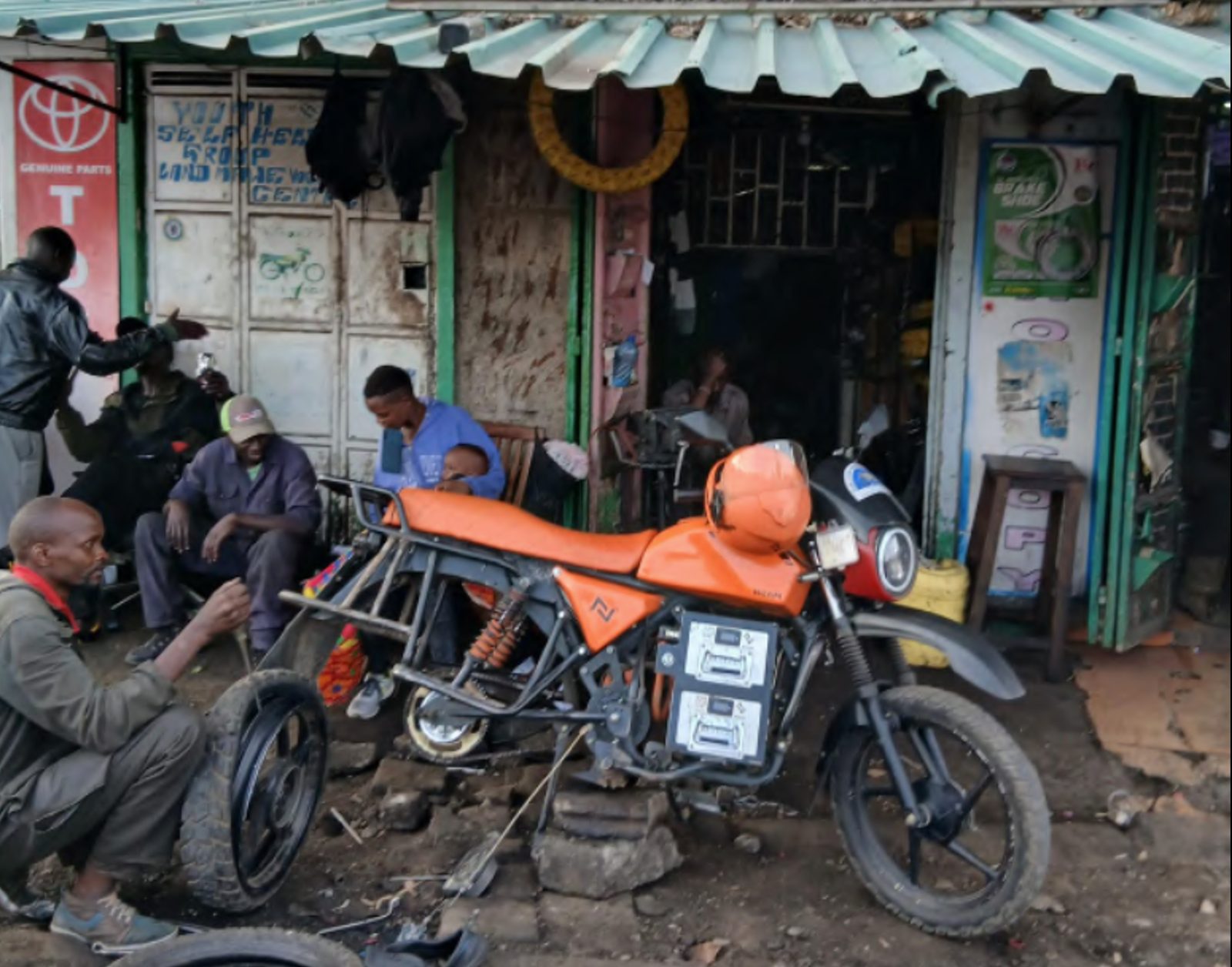 A bright-orange electric motorcycle parked at a small Nairobi hardware kiosk, twin battery packs visible under the seat.