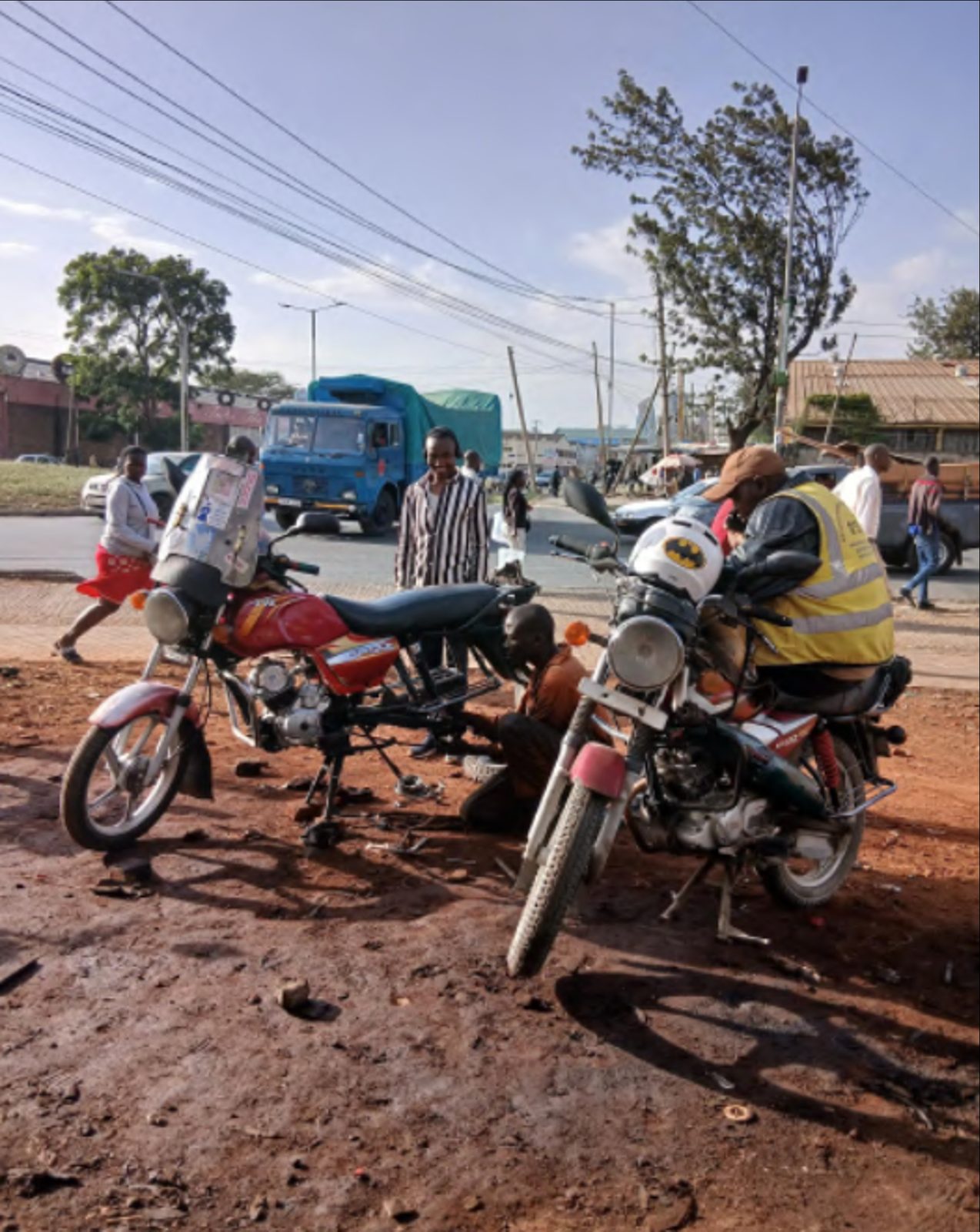 A Nairobi roadside scene at first light: a red Boxer 100 boda-boda propped on its side stand next to a green-and-red boda-boda, a mechanic in an orange top squatting between them working on a chain, another rider in a high-vis vest and a helmet leaning over one of the bikes; a blue lorry in the background, dusty red-earth ground and a busy junction beyond.
