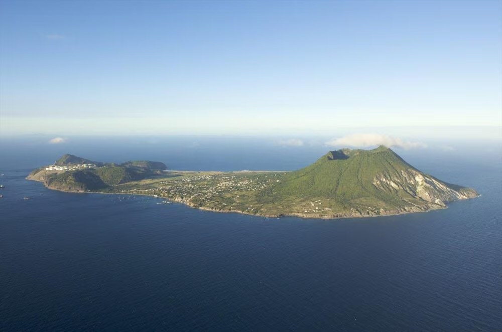Aerial view of St. Eustatius: a small volcanic island with the Quill cone rising to the right, the settled plain and airstrip running across the middle, and industrial oil terminals visible on the low western tip; deep-blue Caribbean sea on all sides.
