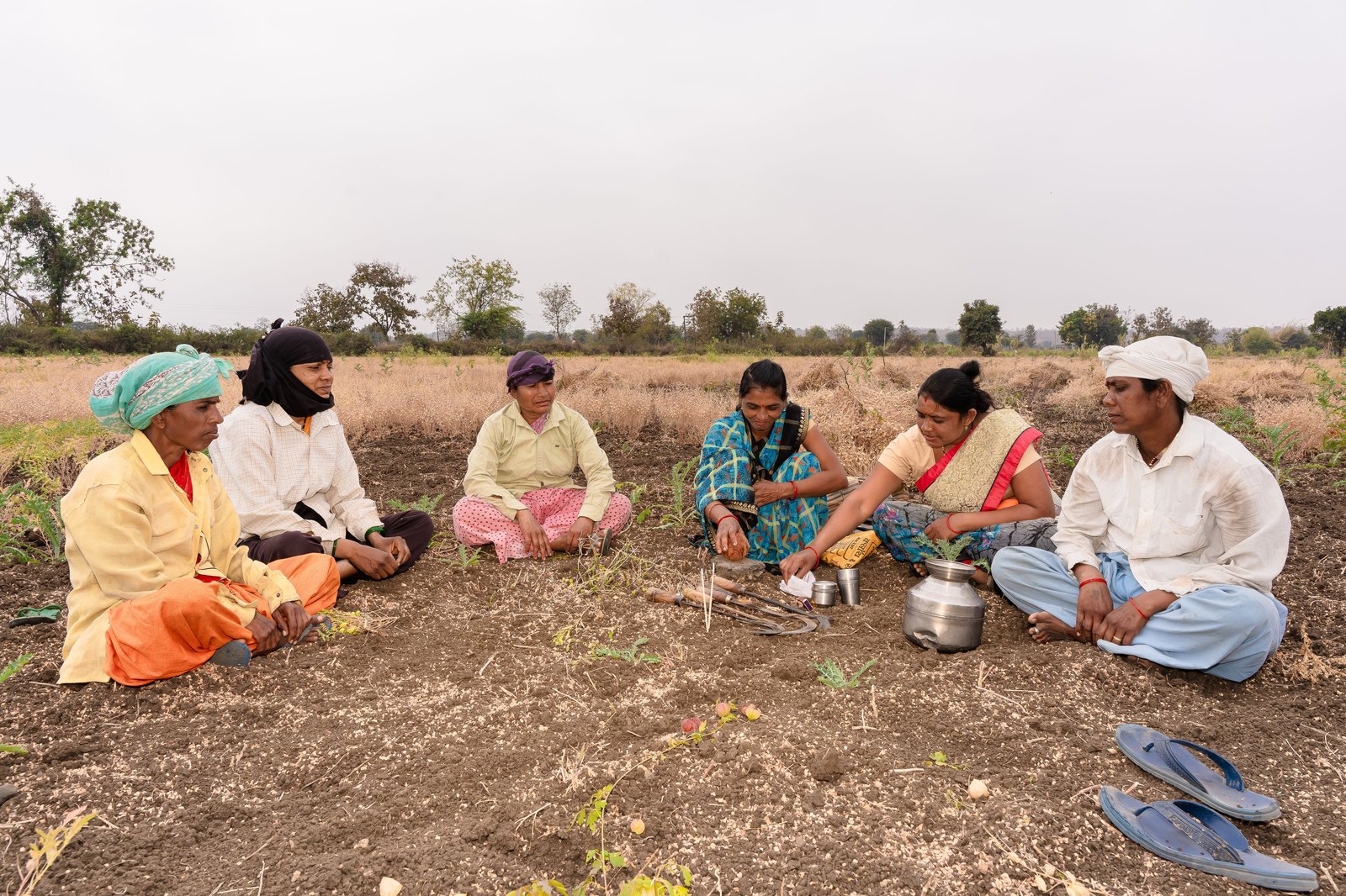 Six women farmers sitting in a circle at the edge of a harvested field, with hand tools and a water pot between them, in conversation.