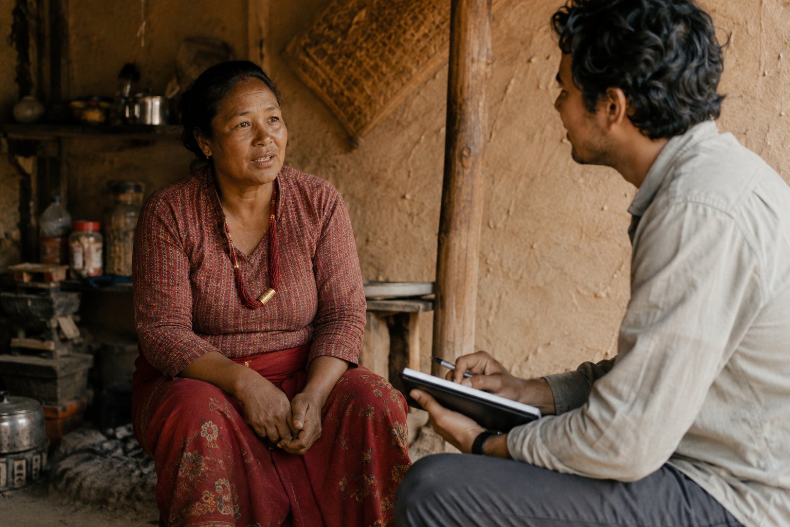 A researcher takes notes while a woman speaks, seated in the doorway of her home. A recorded, consented interview in the setting the respondent lives in.